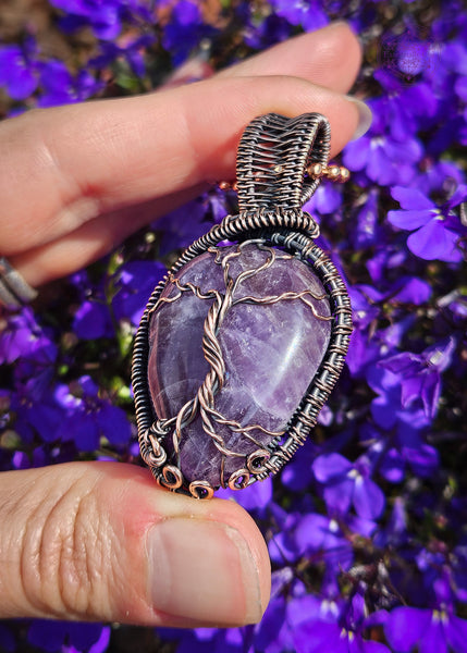 Showing the bottom portion of this copper wire wrapped amethyst tree of life pendant held in hand with purple lobelia flowers in the background. The Amethyst is a dark purple color and the roots have a swirl pattern near the bottom to symbolize the roots growing on rocks. There's a woven pattern along the sides of the pendant and a sturdy bail. 