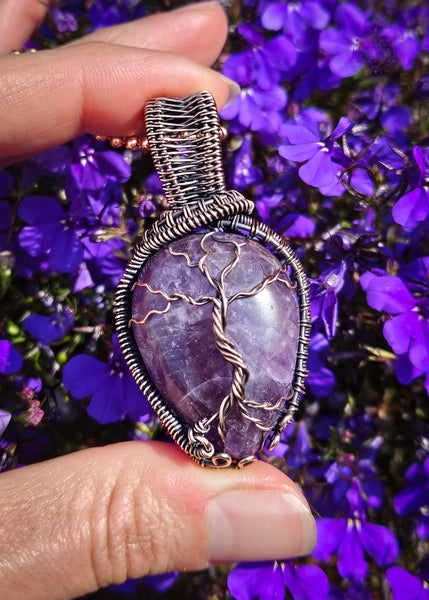 copper wire wrapped amethyst tree of life pendant held in hand with purple lobelia flowers in the background. The Amethyst is a dark purple color, and the roots have a swirl pattern near the bottom to symbolize the roots growing on rocks. There's a woven pattern along the sides of the pendant and a sturdy bail. Shown in full sun to see the white come through on this chevron amethyst.