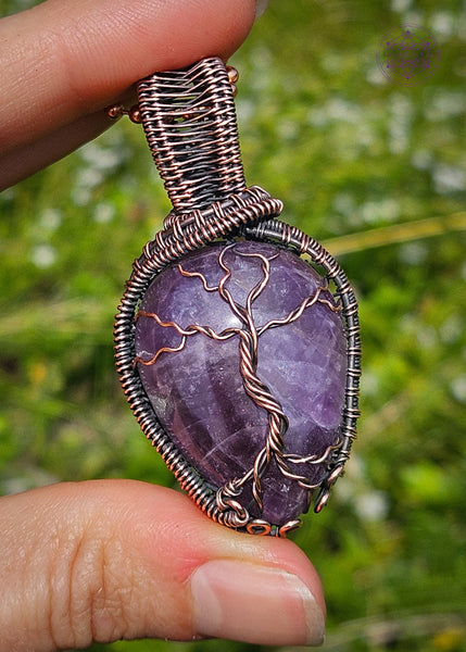 copper wire wrapped amethyst tree of life pendant held in hand with green grass in the background. The Amethyst is a dark purple color and the roots have a swirl pattern near the bottom to symbolize the roots growing on rocks. There's a woven pattern along the sides of the pendant and a sturdy bail. 