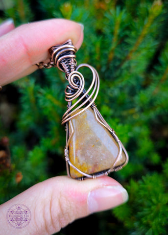 Nebu - a yellow - Orange Aventurine Copper Wire Wrapped Necklace - A captivating triangle-shaped pendant inspired by ancient Egyptian motifs, featuring a three-swoop bail design. The orange aventurine sparkles with golden hues, reminiscent of a radiant sunset. Shown held in hand with a dark evergreen bush in the background to show contrast and details.