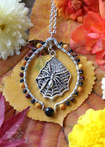 Silver Beaded Hoop Necklace with a spider on a web charm and a faceted black tourmaline focal bead. The necklace is beaded with a gradient of orange, blood red, and matte black glass beads. A stylish and mystical accessory, perfect for adding a touch of gothic and witchy flair to your outfit.