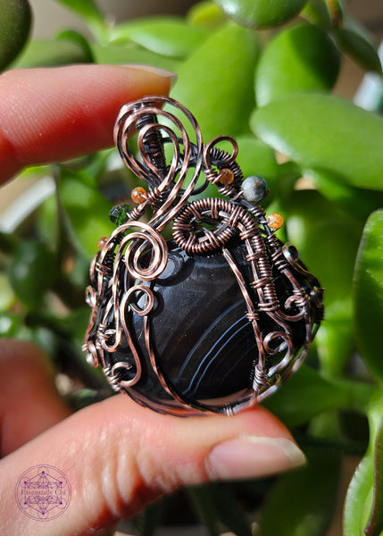 Black agate pumpkin necklace with a copper wire wrapped filagree design and accents of green tourmaline and sunstone crystals in the vines. The black agate crystal has a V shape white and slightly charcoal grey banding with the majority being black.  