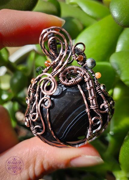 Black agate pumpkin necklace with a copper wire wrapped filagree design and accents of green tourmaline and sunstone crystals in the vines. The black agate crystal has a V shape white and slightly charcoal grey banding with the majority being black.  