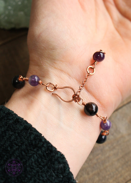 Copper wire wrapped bracelet features 8mm black tourmaline beads individually wrapped with 6mm amethyst beads individually wrapped linking together for an elegant bracelet. Showing the backside of bracelet on wrist to see the hook and the dangle charm. 