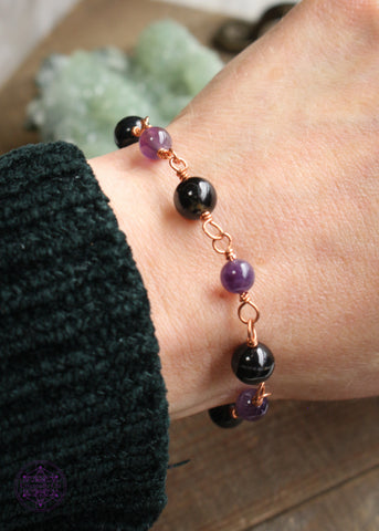 Copper wire wrapped bracelet features 8mm black tourmaline beads individually wrapped with 6mm amethyst beads individually wrapped linking together for an elegant bracelet. Shown on wrist to see how it lays. Green crystals in the background. 