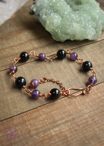 Copper wire wrapped bracelet features 8mm black tourmaline beads individually wrapped with 6mm amethyst beads individually wrapped linking together for an elegant bracelet. A copper hook clasp and an amethyst charm bead dangles at the end. Shown on a wooden board for full preview of all links. Green crystals in the background. 