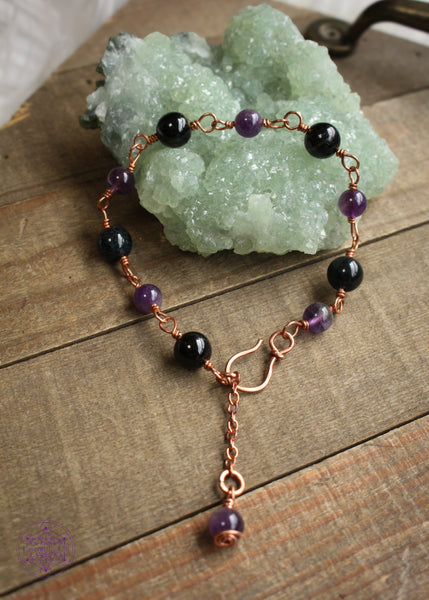 Copper wire wrapped bracelet features 8mm black tourmaline beads individually wrapped with 6mm amethyst beads individually wrapped linking together for an elegant bracelet. Showing the whole bracelet with hook claps and dangle charm laying on a green crystal on a wooden board.