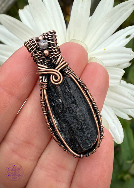 A raw faced black tourmaline oval crystal set in a woven copper wire design. The Sides of the pendant are like a woven basket, the base of the bail has a 180 swoop and the bail has two small copper beads with a smoky quartz between them. Held in hand to show size reference and a white oxeye daisy behind it to make the black pop out. 
