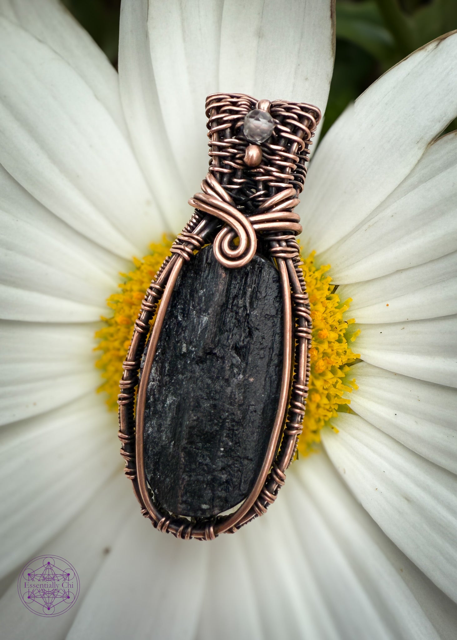 A raw faced black tourmaline oval crystal set in a woven copper wire design. The Sides of the pendant are like a woven basket, the base of the bail has a 180 swoop and the bail has two small copper beads with a smoky quartz between them. Shown sitting on top of a white oxeye daisy to make the black crystal pop out. 