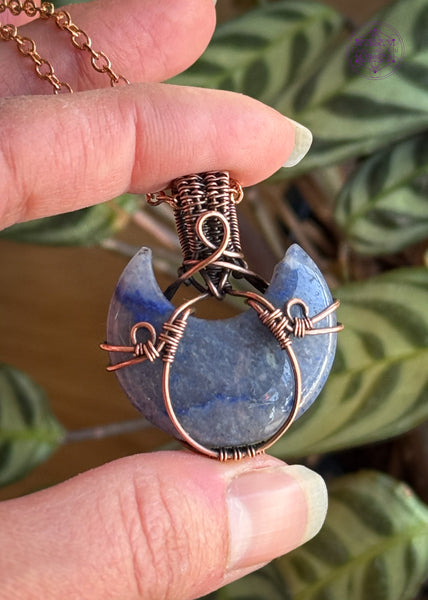 Copper wire wrapped blue aventurine half moon with a simple copper wire wrapped design. Held in hand with plants in the background.