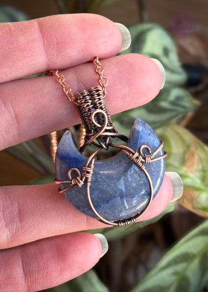 Copper wire wrapped blue aventurine half moon with a simple copper wire wrapped design. Held in hand with plants in the background.