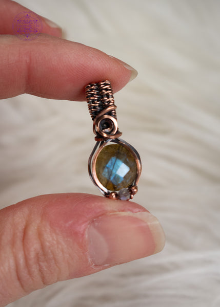 Amazing blue flash labradorite with moonstone copper wire wrapped pendant in a dainty design. The labradorite has a faceted face where it catches the light at every angle and the little moonstone bead at the base of the pendant also has a blue flash. The base of the bail has a small swirl. Held in hand to show the 1" size and a white faux fur background.
