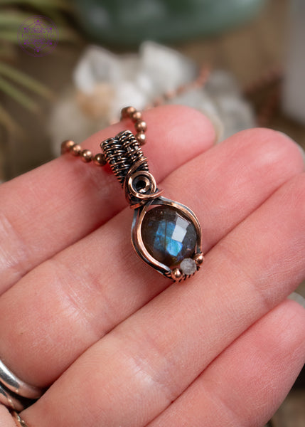 Amazing blue flash labradorite with moonstone copper wire wrapped pendant in a dainty design. The labradorite has a faceted face where it catches the light at every angle and the little moonstone bead at the base of the pendant also has a blue flash. The base of the bail has a small swirl. Held in hand to show dainty size.