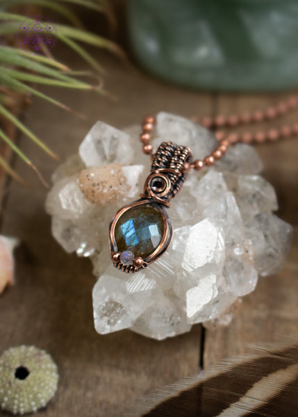 Amazing blue flash labradorite with moonstone copper wire wrapped pendant in a dainty design. The labradorite has a faceted face where it catches the light at every angle, and the little moonstone bead at the base of the pendant also has a blue flash. The base of the bail has a small swirl. Sitting on an apophyllite crystal on a wooden table with the pendant angled to the left.