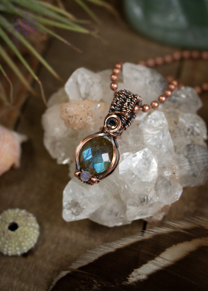 Amazing blue flash labradorite with moonstone copper wire wrapped pendant in a dainty design. The labradorite has a faceted face where it catches the light at every angle, and the little moonstone bead at the base of the pendant also has a blue flash. The base of the bail has a small swirl. Sitting on an apophyllite crystal on a wooden table with the pendant angled to the left.