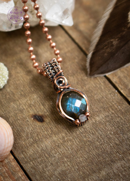Amazing blue flash labradorite with moonstone copper wire wrapped pendant in a dainty design. The labradorite has a faceted face where it catches the light at every angle and the little moonstone bead at the base of the pendant also has a blue flash. The base of the bail has a small swirl. Sitting on a wooden table with a feather and crystals around it.