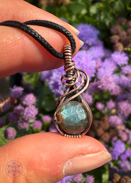 Faceted blue labradorite mini pendant with a subtle swoop from the base of the bail to the right side. The blue flash gets deeper at the bottom of the labradorite like deep water. Meant to look like a raindrop. Held in hand with purple flowers in the background.