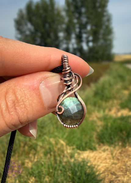 Faceted blue labradorite mini pendant with a subtle swoop from the base of the bail to the right side. The blue flash gets deeper at the bottom of the labradorite like deep water. Meant to look like a raindrop. Held in hand with grassy wetlands in the background.