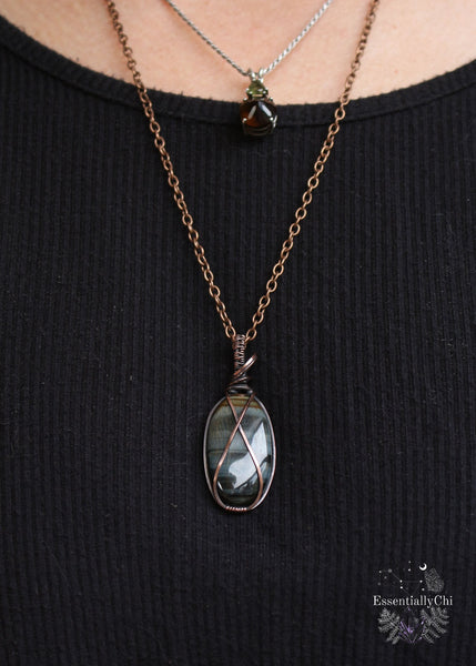Bkue tiger eye oval pendant wrapped in copper wire with a criss cross shape across rhe face of the stone. shown worn around the neck to display how it hangs and the length. 