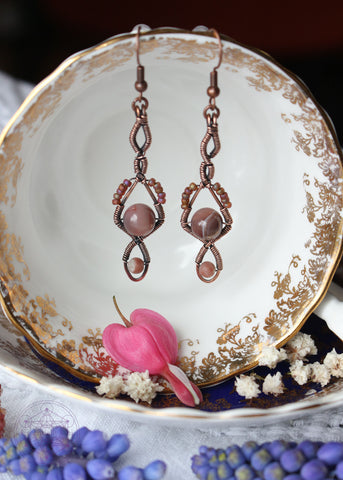 Handcrafted Magnolia earrings featuring rosy pink Botswana agate stones and pink jasper beads wrapped in copper wire. The shape of the earrings are long and narrow with two twists at the top and then a diamond shape with the agate bead in the center and another loop under that with the pink jasper in the center of the loop. Copper wire is antiqued to bring out the details.