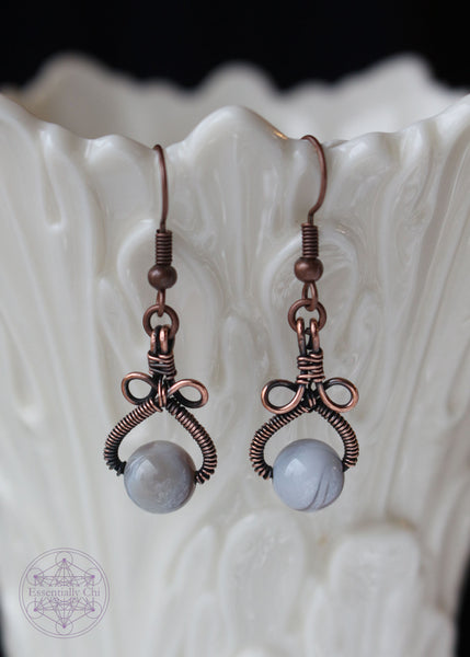 Handcrafted Lily earrings featuring Botswana agate stones wrapped in copper wire. The shape of the earrings is a pear shape with two loops above the stone that looks like a bow. The color of the Botswana agate bead banding is grey and white. Copper wire is antiqued to bring out the details.