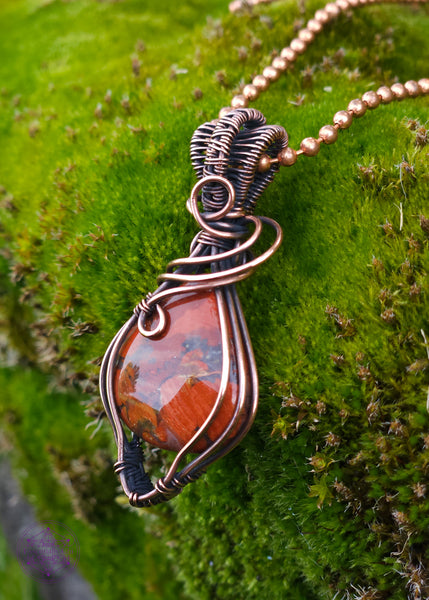 elegant brecciated red jasper pendant wrapped in copper wire. The wire wrap is a tear drop shape downwards, with swirls at the base of the bail. The bail is a double weave style for texture. Displayed on a mossy green background.