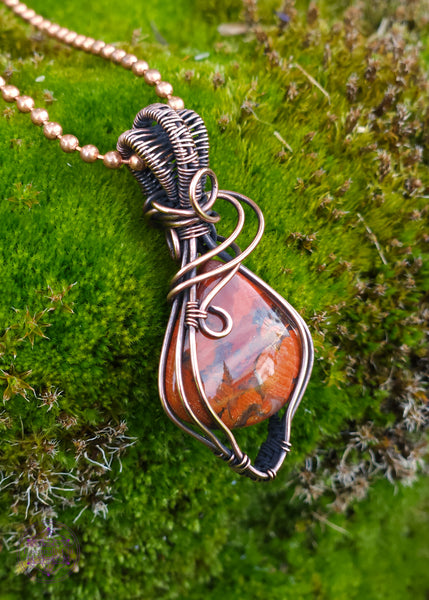 elegant brecciated red jasper pendant wrapped in copper wire. The wire wrap is a tear drop shape downwards, with swirls at the base of the bail. The bail is a double weave style for texture. Displayed on a mossy green background.