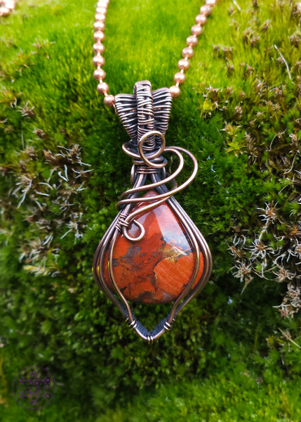 elegant brecciated red jasper pendant wrapped in copper wire. The wire wrap is a tear drop shape downwards, with swirls at the base of the bail. The bail is a double weave style for texture. Displayed on a mossy green background.