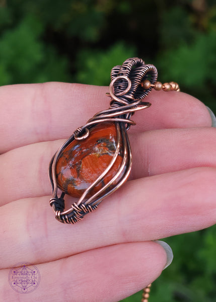 elegant brecciated red jasper pendant wrapped in copper wire. The wire wrap is a tear drop shape downwards, with swirls at the base of the bail. The bail is a double weave style for texture. Held in hand showing the left side of the wrap.