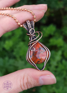 elegant brecciated red jasper pendant wrapped in copper wire. The wire wrap is a tear drop shape downwards, with swirls at the base of the bail. The bail is a double weave style for texture. Shown in hand to show size reference.