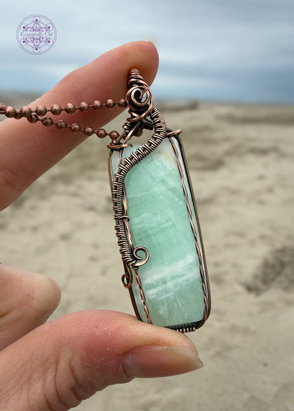A handcrafted copper wire-wrapped pendant with a soft light green Caribbean calcite rectangular stone held by a hand on a beach. The wires form a curl along the left side, simulating a wave crashing into Devil's Churn on the central Oregon coast.