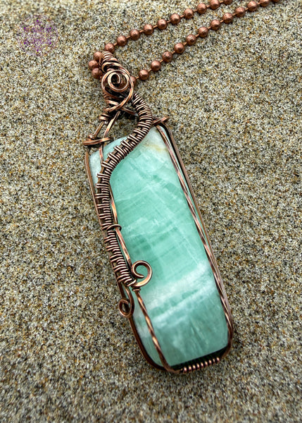 A handcrafted copper wire-wrapped pendant with a soft light green Caribbean calcite rectangular stone shown sitting on the sand for the beachy vibe. The wires form a curl along the left side, simulating a wave crashing into Devil's Churn on the central Oregon coast.