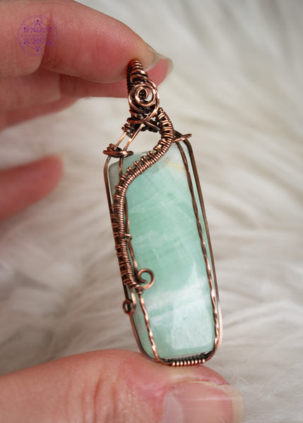 A handcrafted copper wire-wrapped pendant with a soft light green Caribbean calcite rectangular stone held by a hand with a white faux fur background. The wires form a curl along the left side, simulating a wave crashing into Devil's Churn on the central Oregon coast.