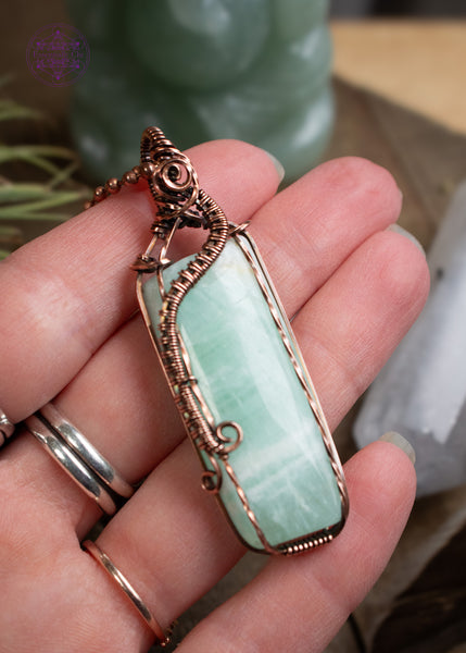 A handcrafted copper wire-wrapped pendant with a soft light green Caribbean calcite rectangular stone held in hand for size reference with crystals in the background. The wires form a curl along the left side, simulating a wave crashing into Devil's Churn on the central Oregon coast.