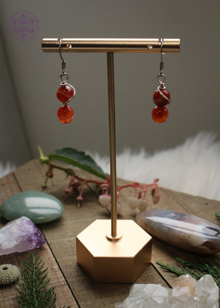 firey red carnelian beaded dangle earrings with a plated silver wire swirled around both beads. Hung on sterling silver hooks. Hanging from earring display to show how they dangle, and how the light shines through them.