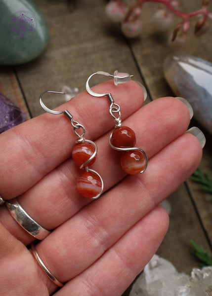 red carnelian beaded dangle earrings with a plated silver wire swirled around both beads. Hung on sterling silver hooks. Held in hand to show size reference.