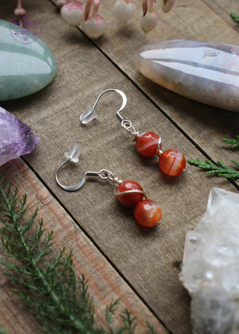 firey red carnelian beaded dangle earrings with a plated silver wire swirled around both beads. Hung on sterling silver hooks. Laying on a wooden table with crystals and foliage around it.