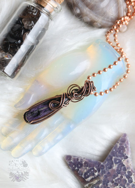 A stunning Charoite Copper Wire Wrapped Necklace in a narrow teardrop shape, expertly adorned with swirling copper wire to accentuate the exquisite charm of the charoite stone. This metaphysical accessory promotes spiritual growth, intuition, and protection, making it a unique and meaningful addition to your jewelry collection.