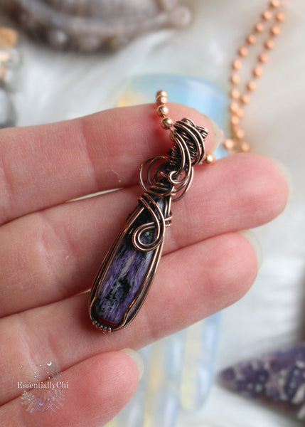 A stunning Charoite Copper Wire Wrapped Necklace in a narrow teardrop shape, expertly adorned with swirling copper wire to accentuate the exquisite charm of the charoite stone. This metaphysical accessory promotes spiritual growth, intuition, and protection, making it a unique and meaningful addition to your jewelry collection.