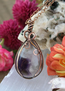 Xenia Chevron Amethyst Pendant with Copper Wire Bow - A symbol of love and gratitude with a range of dark to light purple amethyst. Ideal for Aries, Pisces, and Virgo. Metaphysical properties for intuition, emotional balance, and spiritual growth. Image shown on display with crystals and flowers around it.