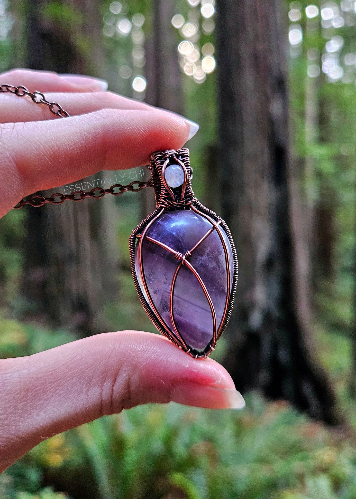 Chevron Amethyst and Rainbow Moonstone copper wire-wrapped jewelry piece with filigree details. Chevron Amethyst criss-cross design on the front, and a blue flash Rainbow Moonstone in the bail. Back features intricate filigree design. Held up showing the redwood forest behind it. 