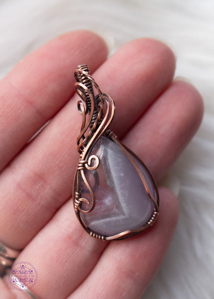 Copper wire wrapped chevron amethyst tear drop shaped crystal shown held in hand for size reference and a white fluffy background. The wires around the bail swoop elegantly down along the right side and end in two small swirls on the left side. he Chevron pattern goes from lavender to a thin band of white and then ends with a pale translucent amethyst.