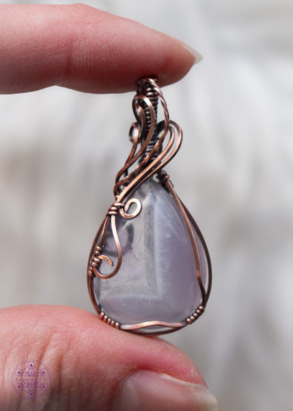 Copper wire wrapped chevron amethyst tear drop shaped crystal shown held in hand with a white fluffy background. The wires around the bail swoop elegantly down along the right side and end in two small swirls on the left side. he Chevron pattern goes from lavender to a thin band of white and then ends with a pale translucent amethyst.