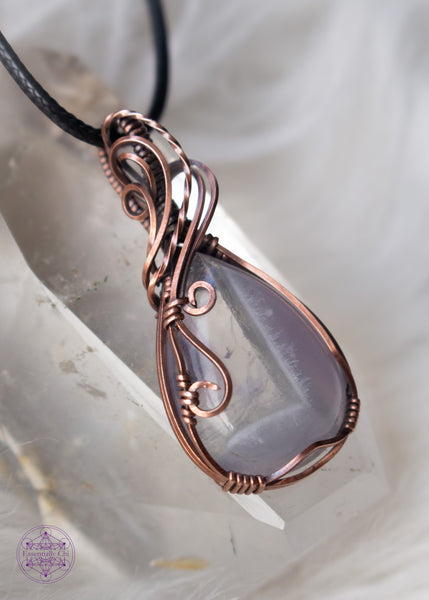 Copper wire wrapped chevron amethyst tear drop shaped crystal sitting on a clear quartz tower with a white fluffy background. The wires around the bail swoop elegantly down along the right side and end in two small swirls on the left side. he Chevron pattern goes from lavender to a thin band of white and then ends with a pale translucent amethyst with a rainbow at the top.