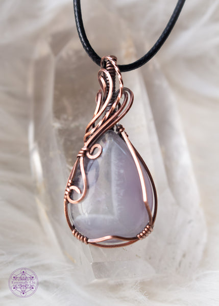 Copper wire wrapped chevron amethyst tear drop shaped crystal shown over a clear quartz tower from the front view and a white fluffy background. The wires around the bail swoop elegantly down along the right side and end in two small swirls on the left side. he Chevron pattern goes from lavender to a thin band of white and then ends with a pale translucent amethyst.