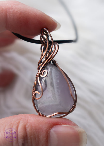 Copper wire wrapped chevron amethyst tear drop shaped crystal shown held in hand with a white fluffy background. The wires around the bail swoop elegantly down along the right side and end in two small swirls on the left side. he Chevron pattern goes from lavender to a thin band of white and then ends with a pale translucent amethyst.