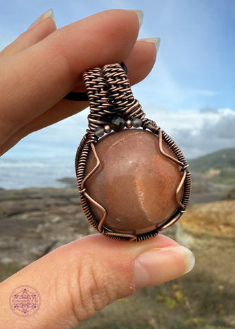 Crystal ball necklace with a chocolate agate crystal that acts as a fidgit spinner and has some small crystal beads of smoky quartz and tourmaline for extra grounding and a little sparkle. Shown held in hand with the blue sky and ocean behind it. 
