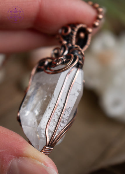 Clear quartz pendant with filigree swirls along the top and side, held in hand with airplants behind it. The angel shows off the record keepers along the right side of the crystal.