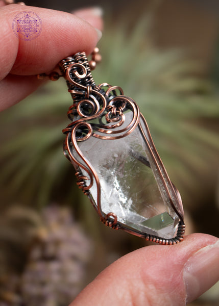 Clear quartz pendant with filigree swirls along the top and side, held in hand with airplants behind it. The angel shows off the clarity of the crystal.