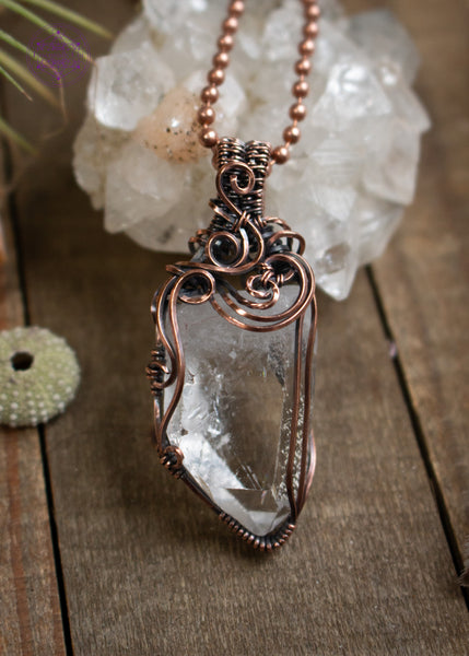 Clear quartz pendant with filigree swirls along the top and side, sitting on a wooden board with crystals and shells around it. The angel shows off the swirls on the front.
